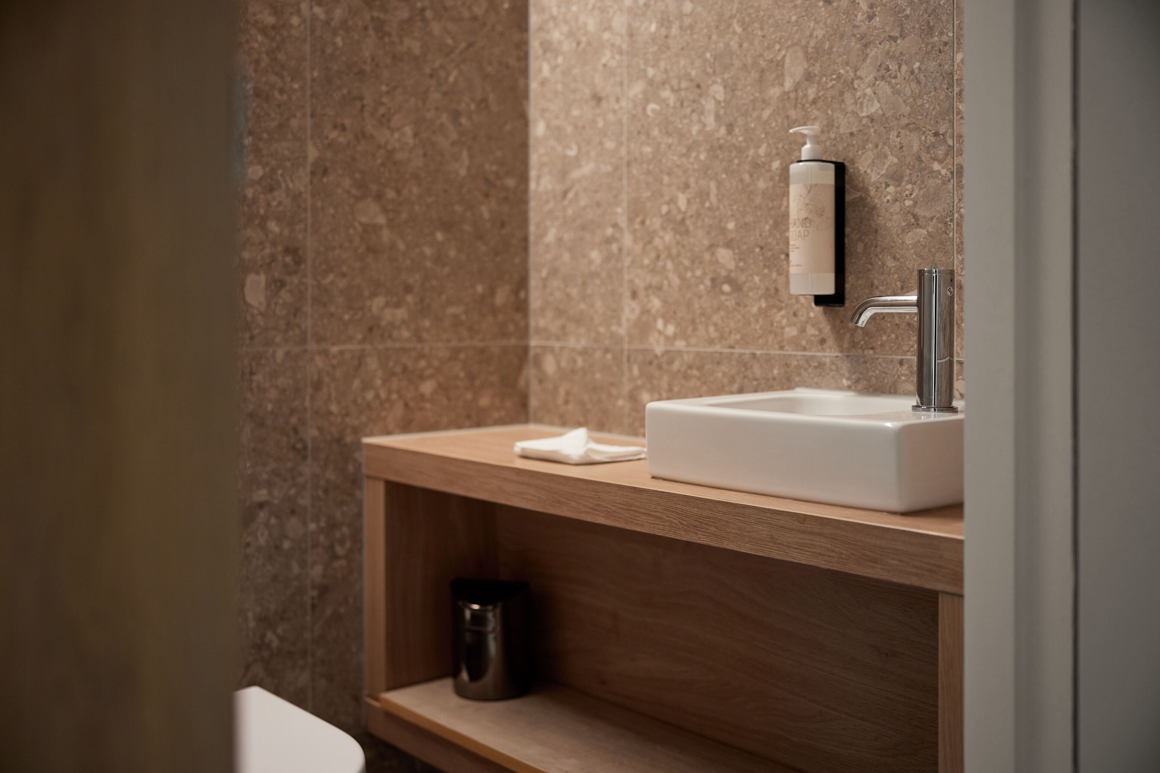 A modern bathroom with a marble wall, a wooden vanity, a white sink, and a soap dispenser.