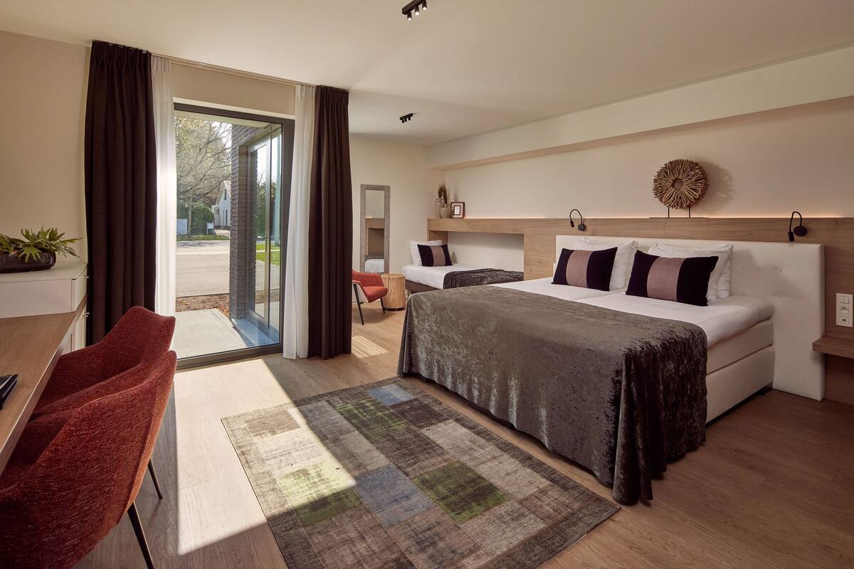 Une chambre d’hôtel moderne, dotée d’un grand lit, d’un bureau et d’une chaise. Une porte vitrée offre une vue sur l’extérieur.