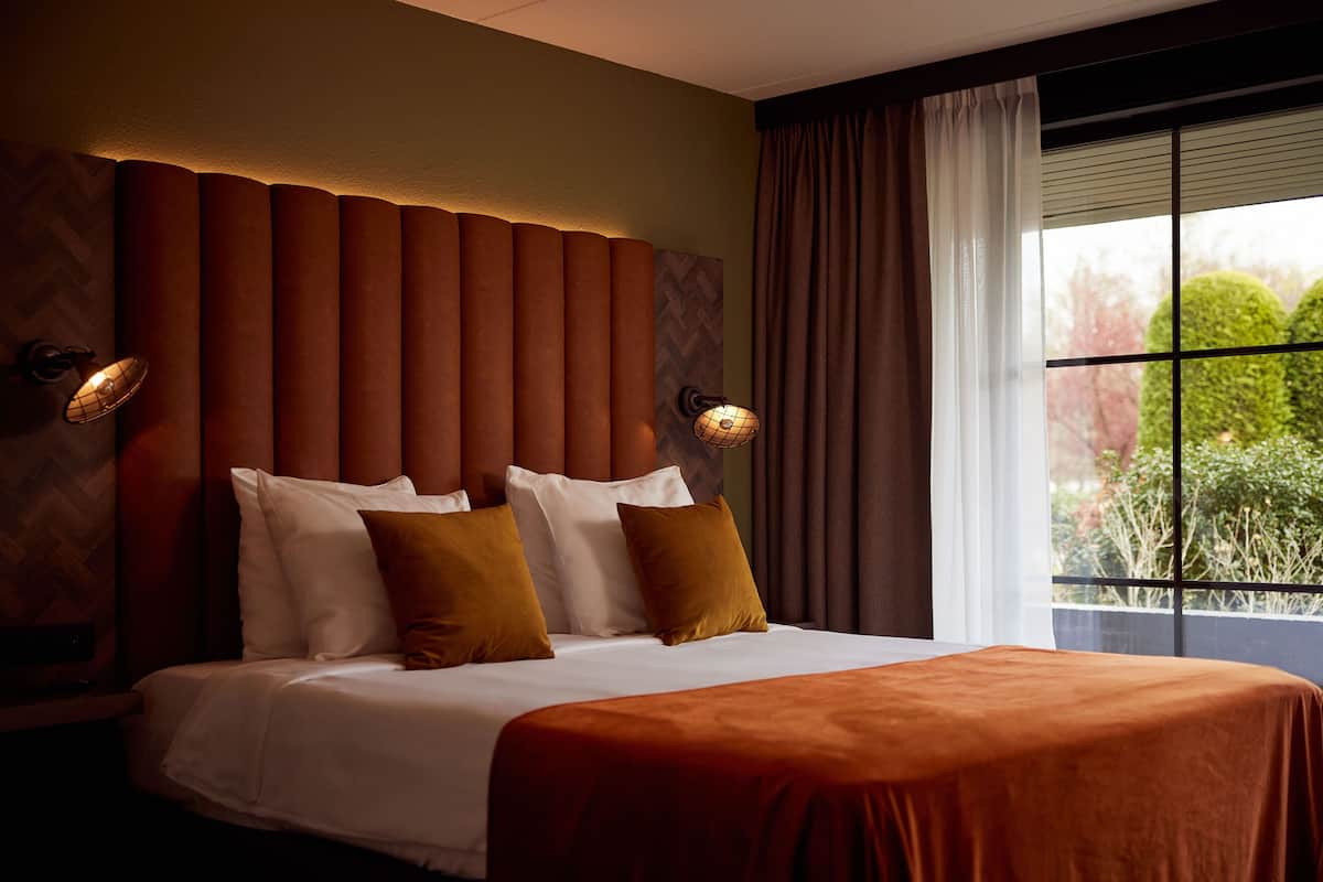 Une chambre d’hôtel avec un grand lit, une tête de lit à lattes verticales, deux appliques murales et une fenêtre avec des rideaux.