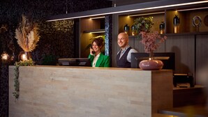 A reception desk with a wooden counter, a potted plant, and a vase of flowers. - Van Der Valk Hotel Dennenhof (Brasschaat)
