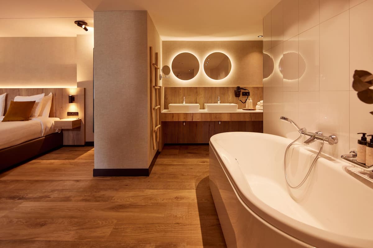 Une salle de bain d’hôtel moderne, dotée d’une grande baignoire, de deux lavabos et d’un miroir avec des lumières disposées en cercle.