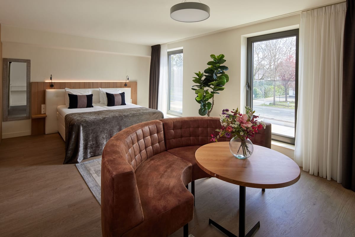 Une chambre d’hôtel moderne, équipée d’un lit, d’un coin salon, d’une table agrémentée d’un vase de fleurs et d’une grande fenêtre avec des rideaux.