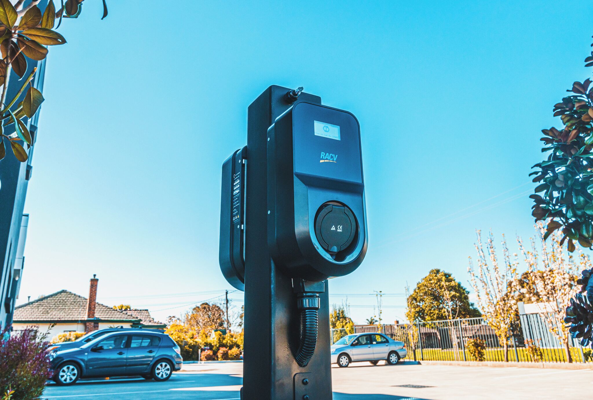 A parking meter with a digital display, located in a residential area with houses and cars in the background.