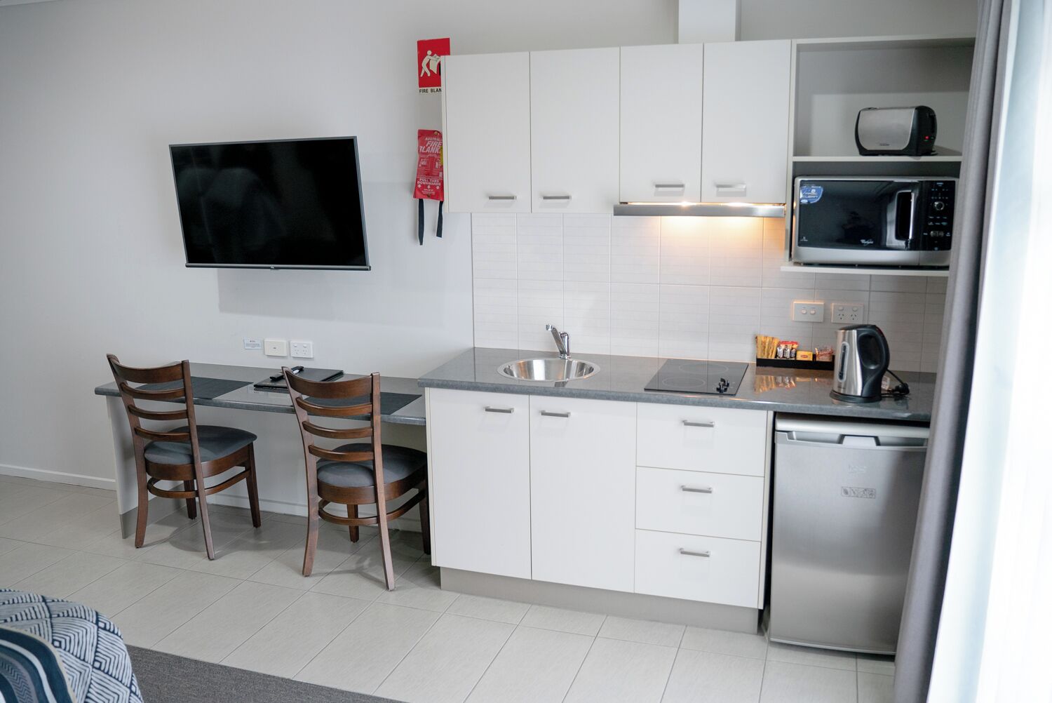 A modern kitchen with white cabinets, a stainless steel sink, a microwave, and a toaster. There is a flat-screen TV mounted on the wall and two wooden chairs at the dining table.
