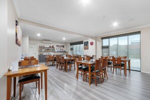 A dining area with multiple wooden tables and chairs, a kitchen in the background, and large windows. - Bridges on Argyle Motel & Apartments (Traralgon)
