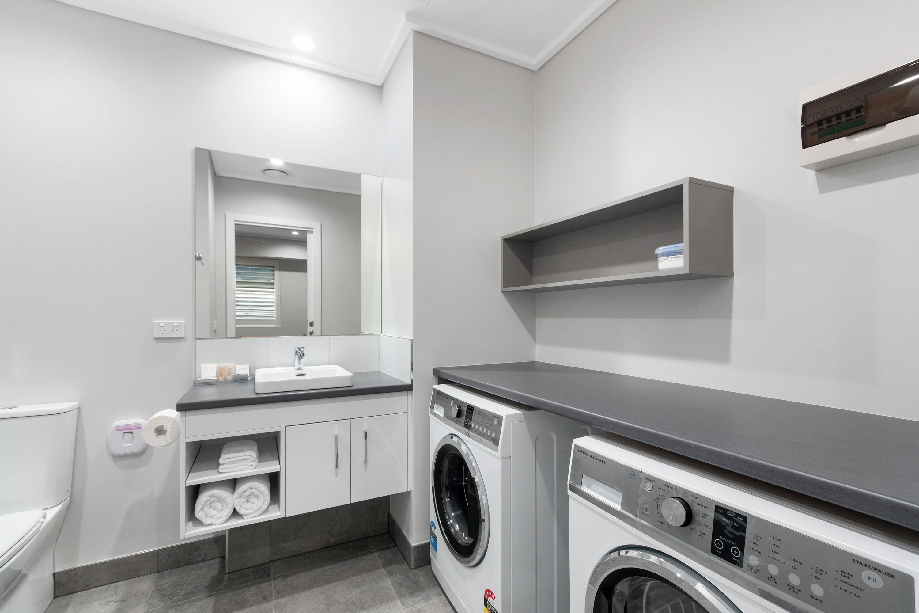 Een moderne badkamer met toilet, wastafel, spiegel, planken en een wasmachine.