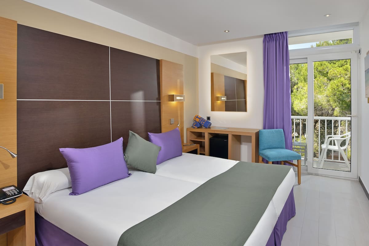 Quarto de hotel com cama, escrivaninha, cadeira e varanda com vista para árvores.