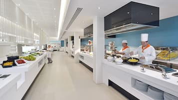 A modern kitchen with chefs preparing food, white countertops, and a variety of food items displayed.