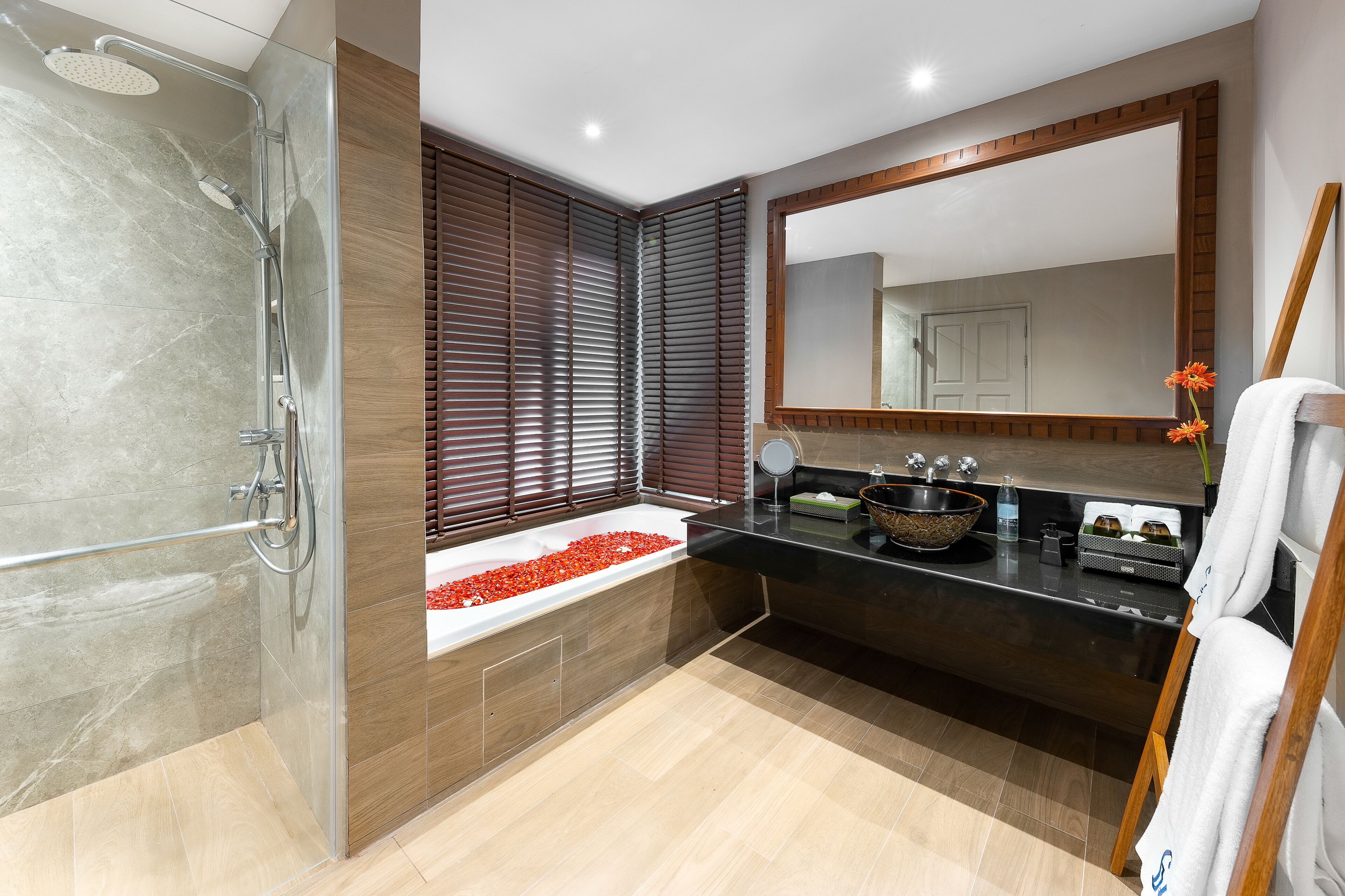 A modern bathroom with a glass-enclosed shower, a bathtub with red pebbles, a large mirror, and a wooden vanity.