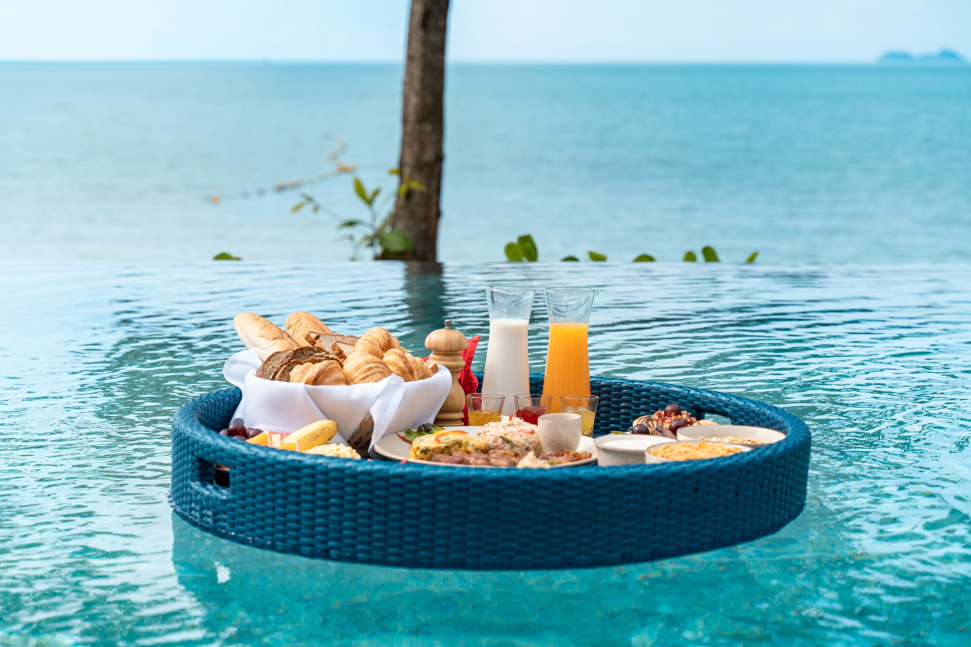 A floating breakfast tray with croissants, pastries, and beverages in a clear blue pool.