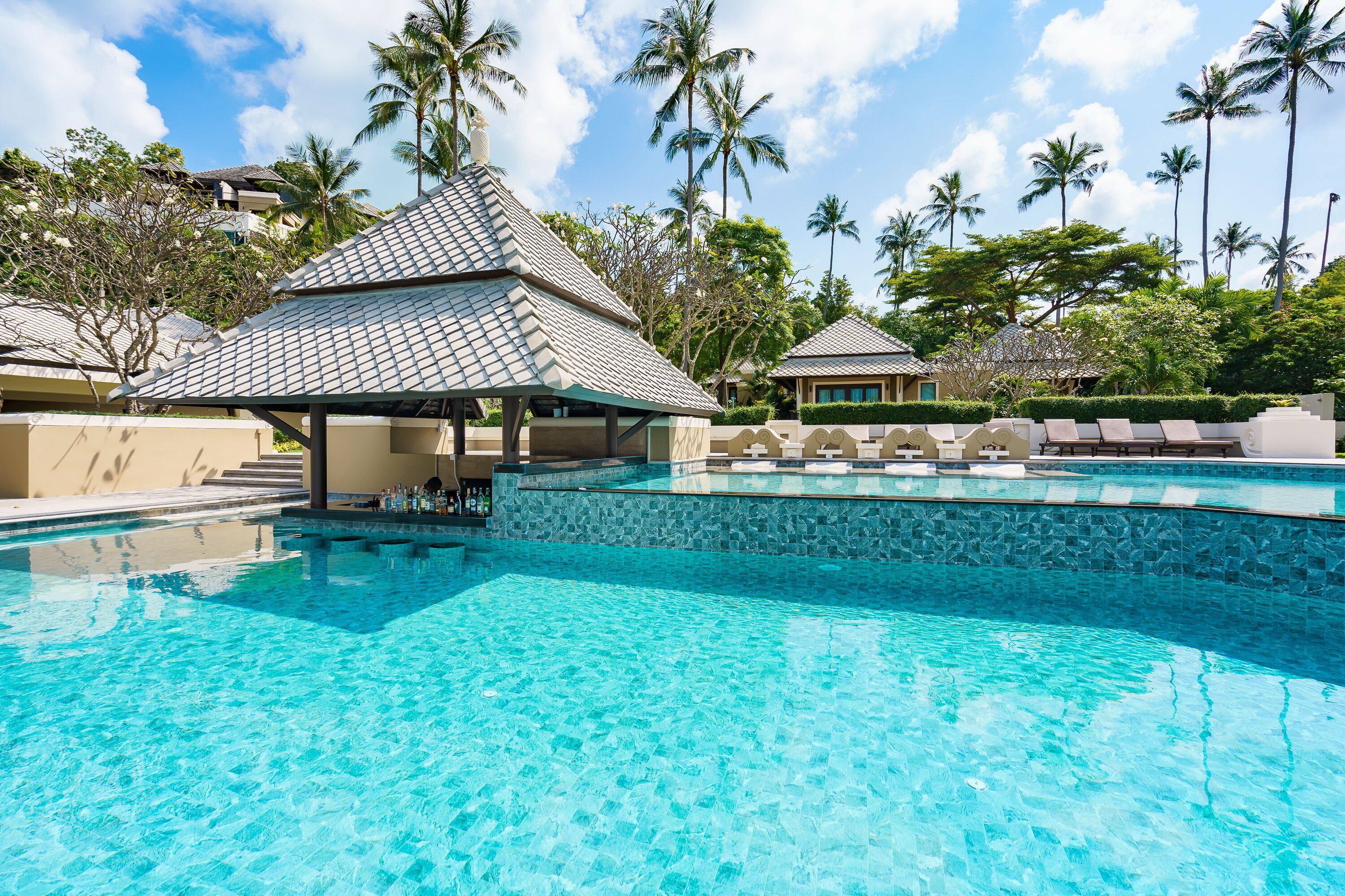 Une piscine d’hôtel avec une eau cristalline, entourée de palmiers et dotée d’un espace détente abrité.