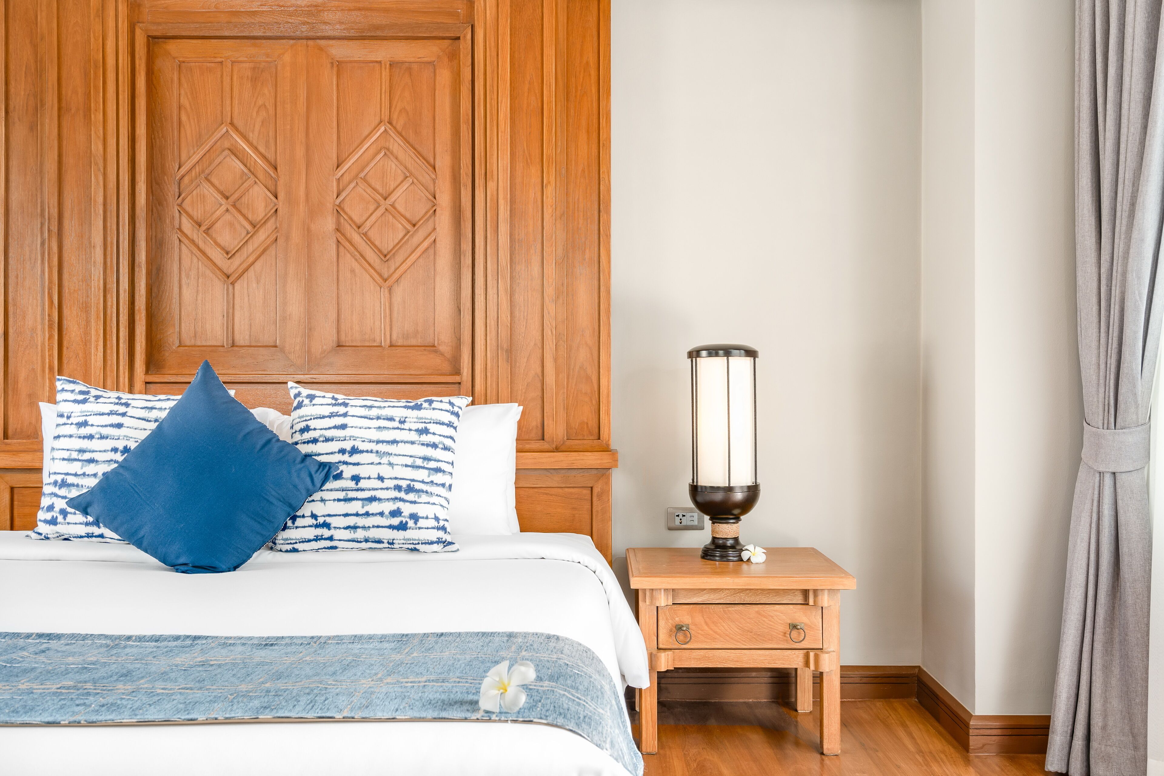 Una habitación de hotel con cabecera de madera, cama con almohadas de diseño azul y blanco, mesita de noche con lámpara y mesita auxiliar de madera.