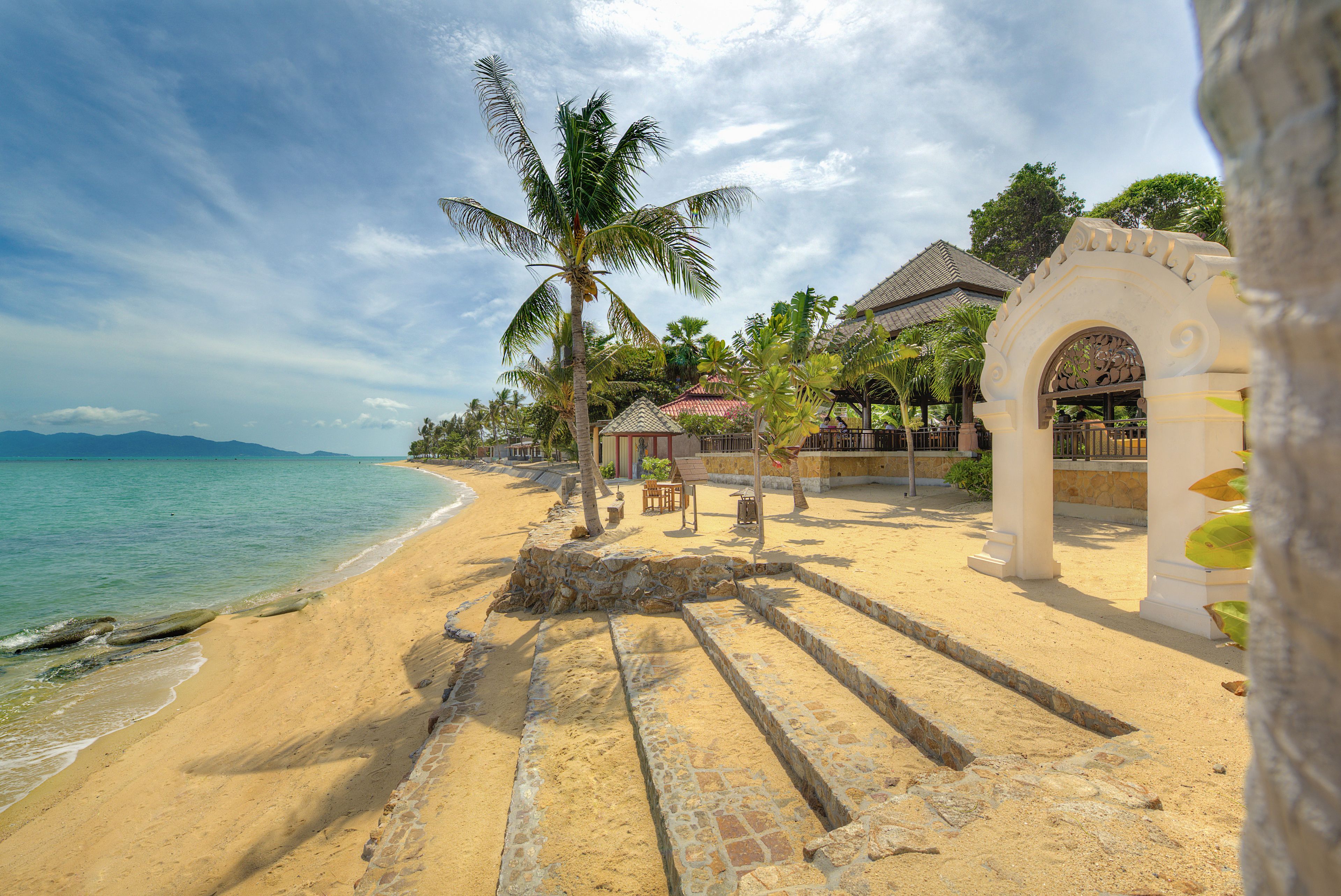 A beach with a stone pathway leading to a gazebo, palm trees, and a building with a tiled roof.