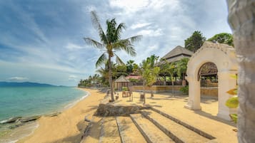 A beach with a stone pathway leading to a gazebo, palm trees, and a building with a tiled roof.
