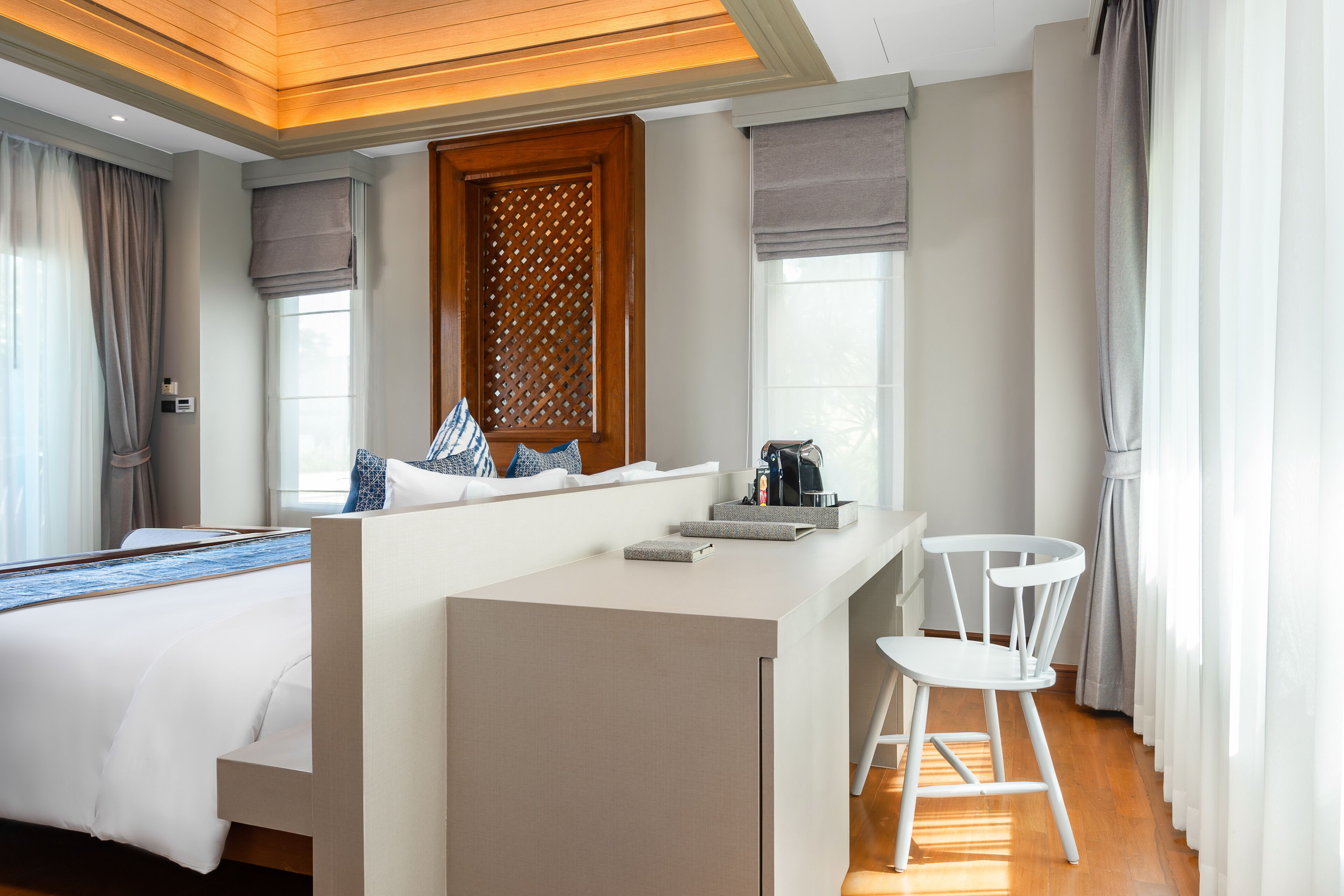 A modern hotel room with a bed, a small kitchenette, a white chair, and a large window with curtains.