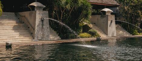 A pool with a stone staircase leading to a covered area, surrounded by lush greenery.