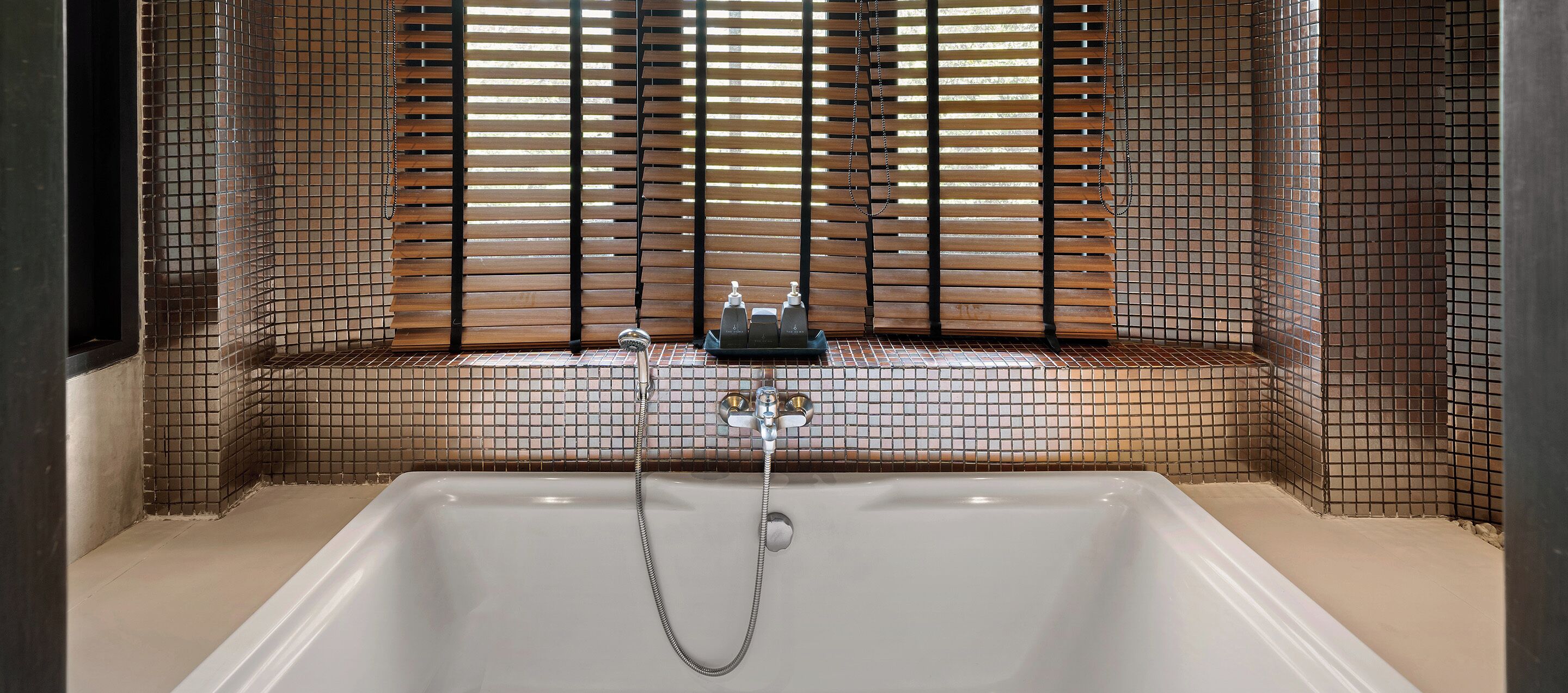 A bathtub with a showerhead and faucet, a window with blinds, and a tiled wall.