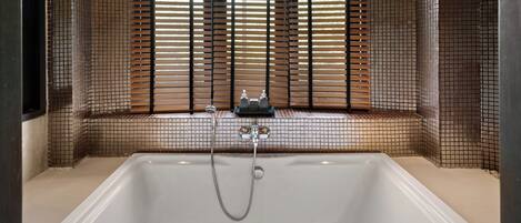 A bathtub with a showerhead and faucet, a window with blinds, and a tiled wall.