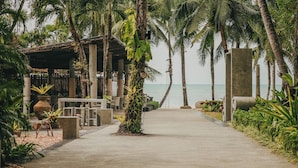 A pathway leading to a beachside area with palm trees, outdoor seating, and a view of the ocean. - The Dewa Koh Chang (Ko Chang)