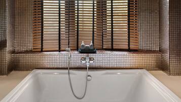 A bathtub with a showerhead, a window with blinds, and a tiled wall.