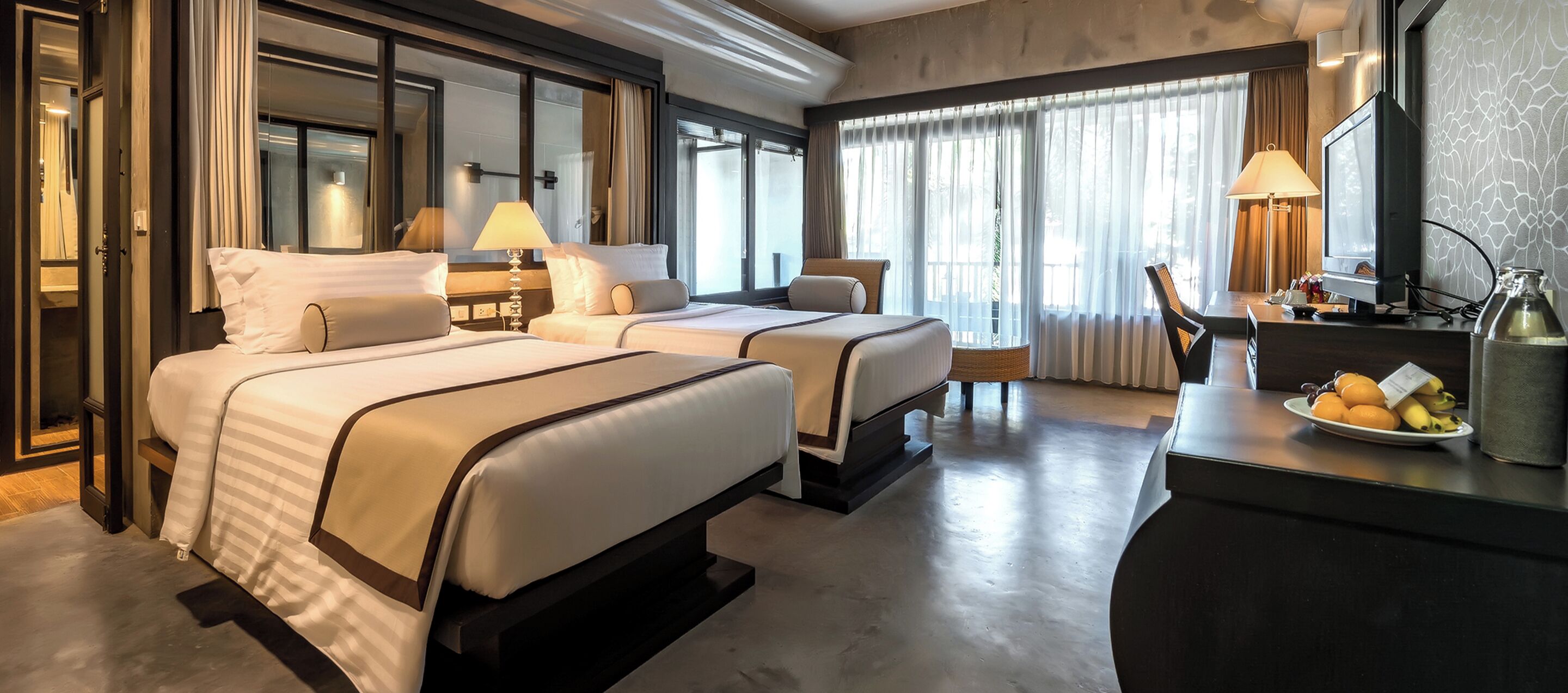 A hotel room with two beds, a desk with a computer, a chair, and a dining table with fruit.