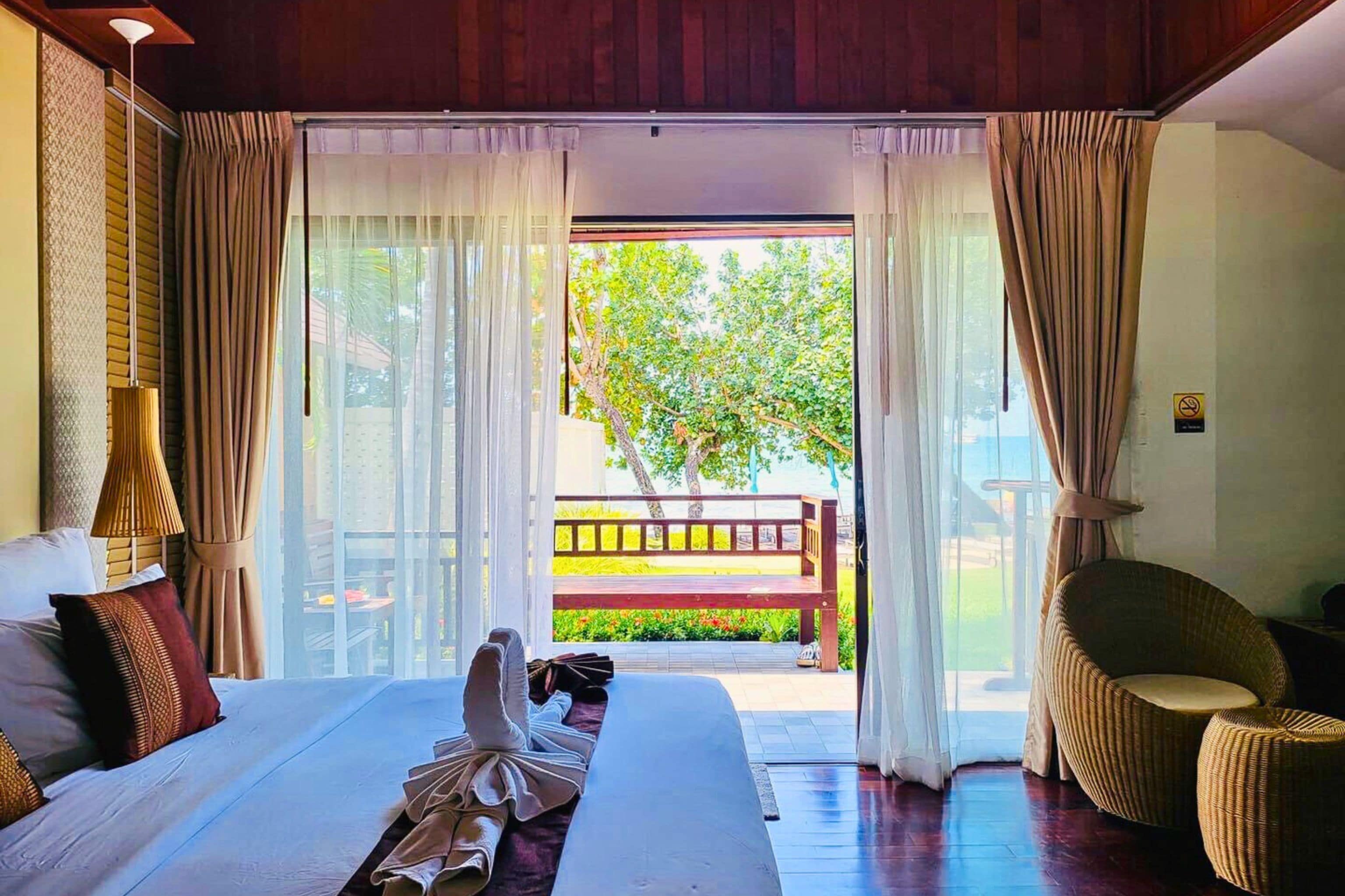 A hotel room with a large bed, a wicker chair, and a view of a garden through a glass door.