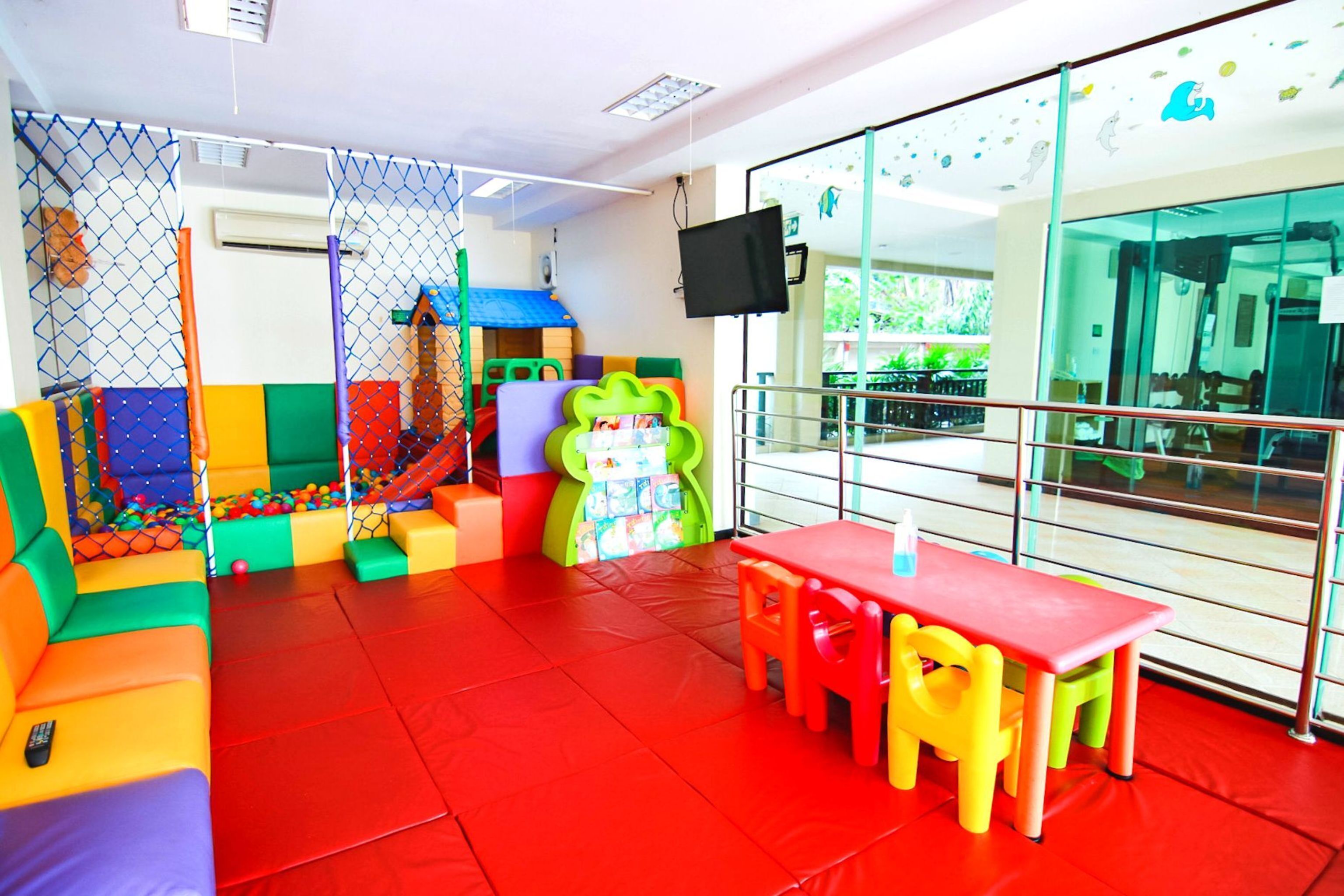 A children's play area with colorful seating and play structures, a red floor, and a glass partition with a view of the outside.