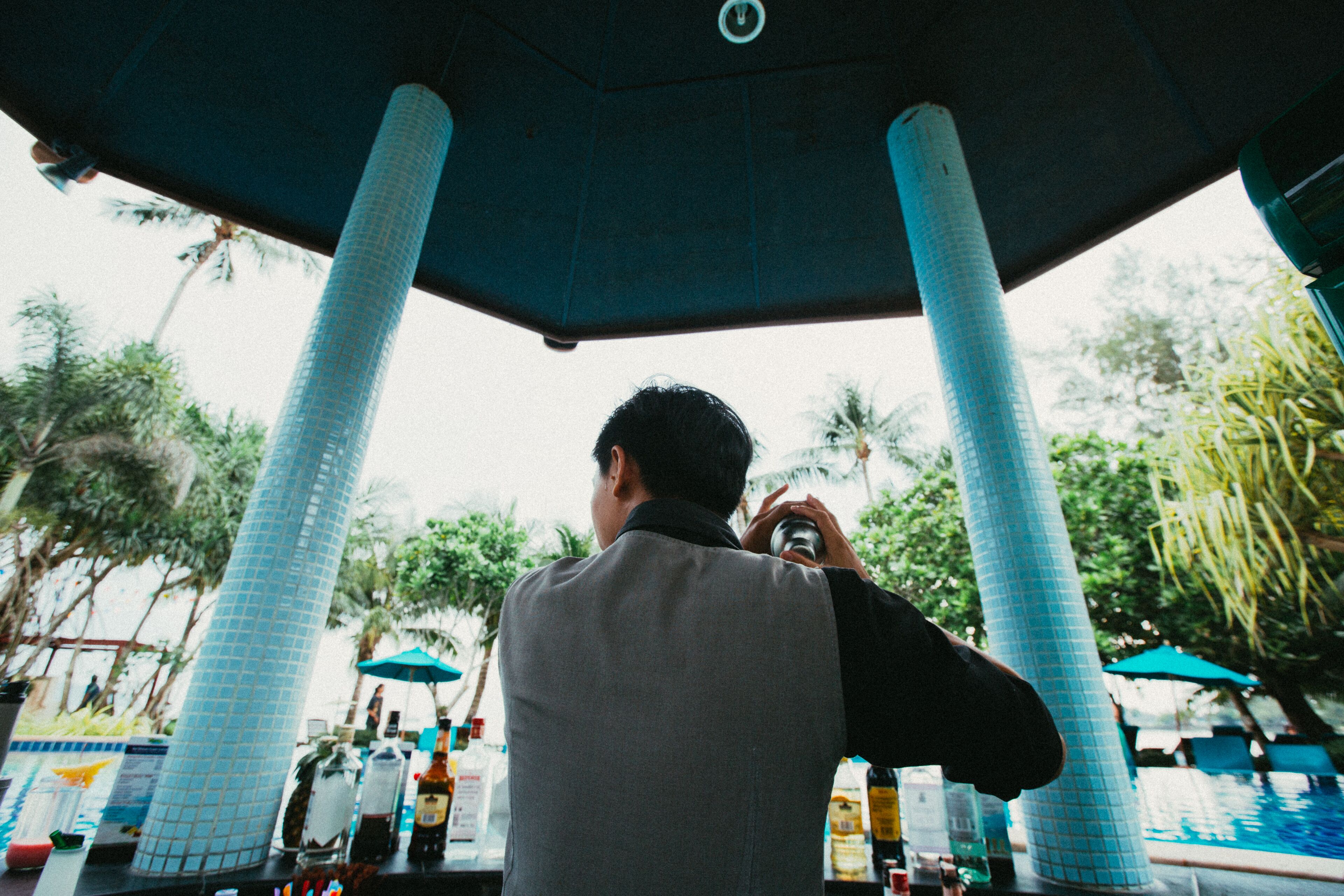 A person taking a photo under a tiled structure with a pool and palm trees in the background.