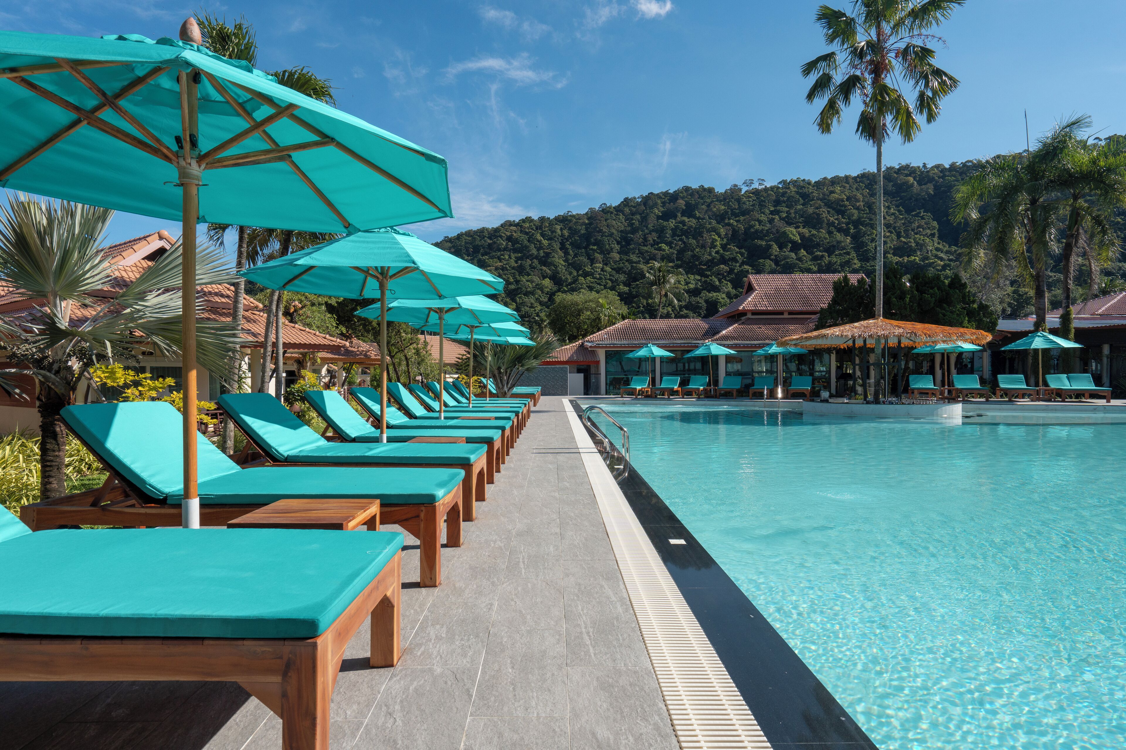 A poolside area with lounge chairs and umbrellas, a clear pool, and a mountainous backdrop.
