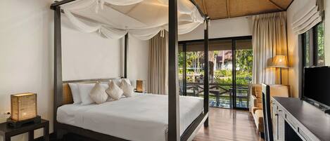 A four-poster bed with white linens, a bedside table with a lamp, and a view of the outdoor garden through a glass door.