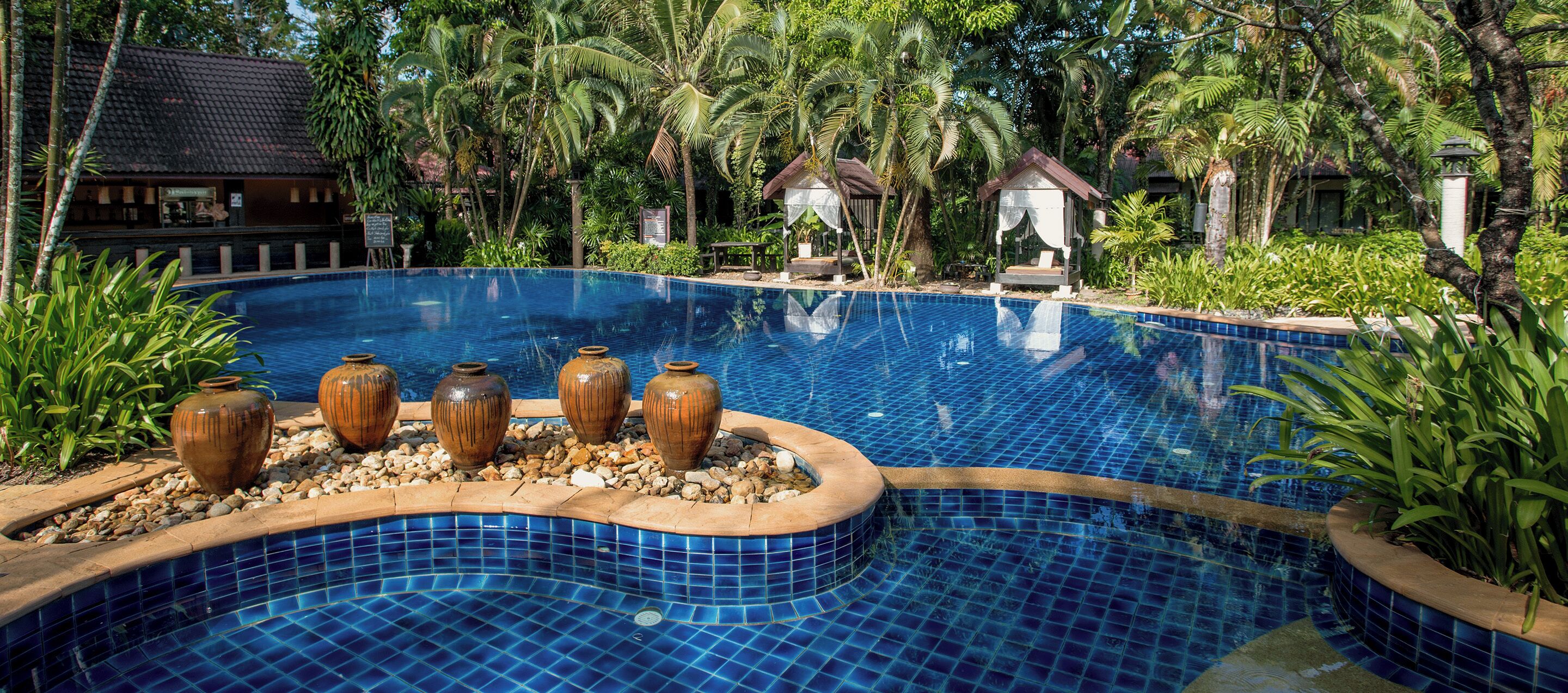 A swimming pool with blue mosaic tiles, surrounded by tropical plants and three large ceramic pots.