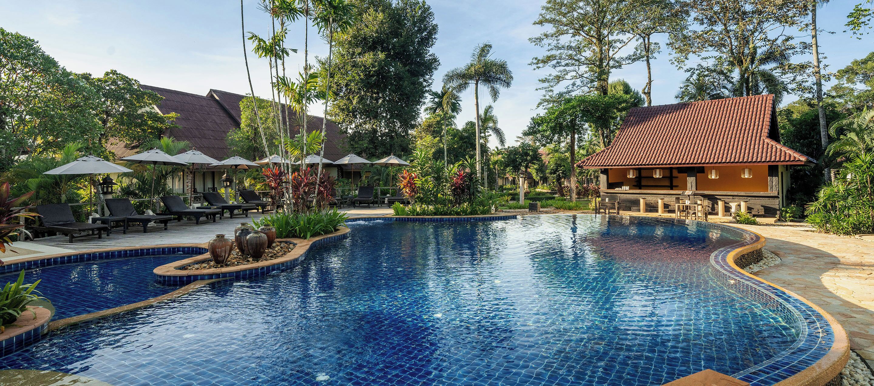 Une piscine entourée de végétation tropicale, agrémentée de chaises longues et de parasols, et bordée d’un bâtiment dont le toit est recouvert de tuiles.