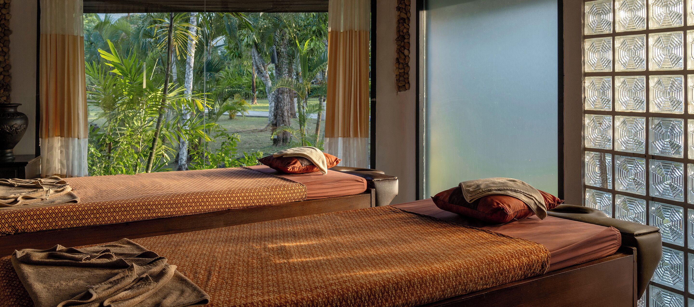 Two massage therapy beds with towels and pillows, a window with a view of greenery, and a decorative wall panel.