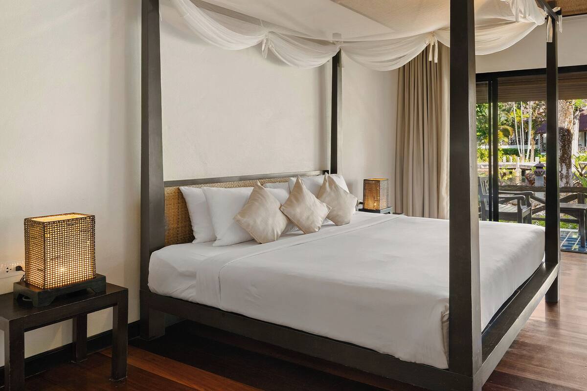A four-poster bed with white linens, a television on a wooden cabinet, and a balcony with outdoor furniture.