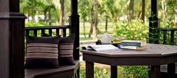 Um banco de madeira com almofadas listradas, uma mesa com chaleira, copos e livros, em uma área externa verde e exuberante.