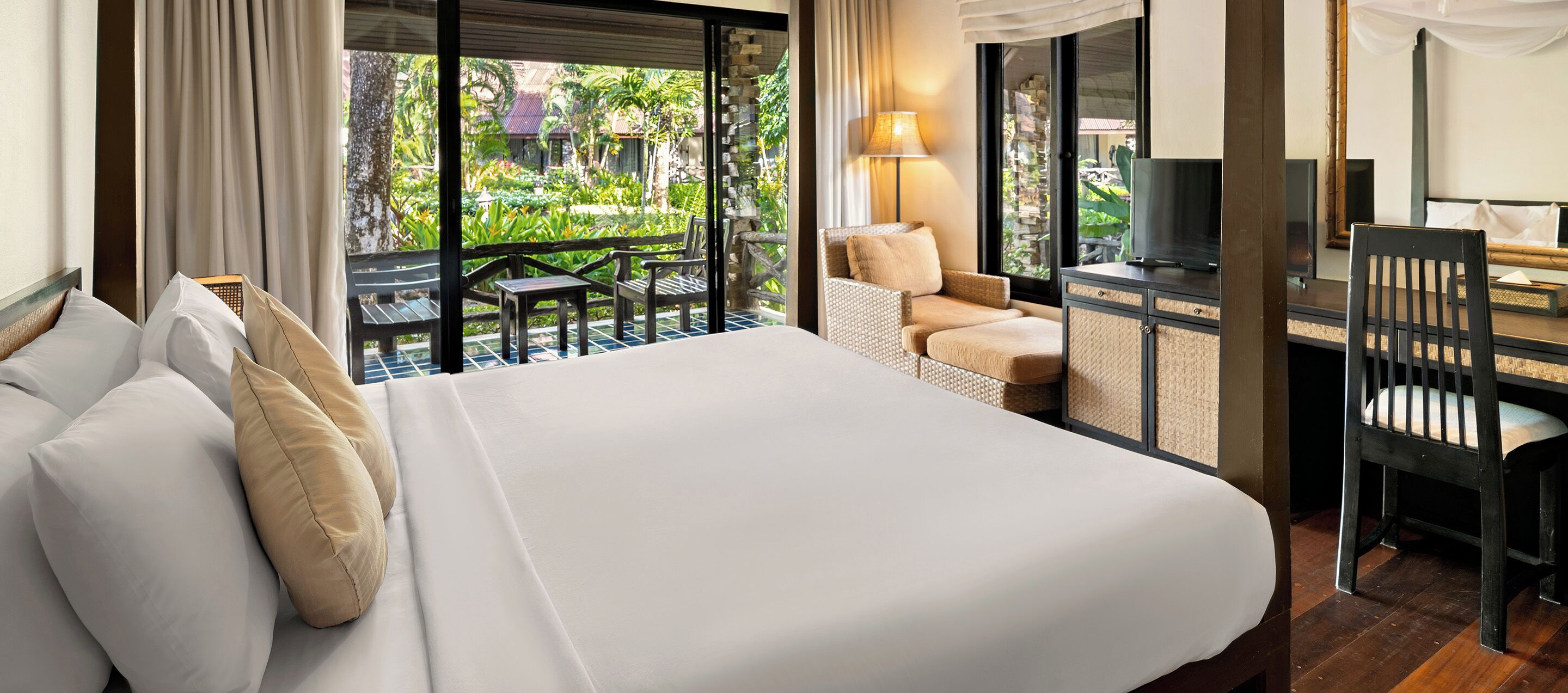 A hotel room with a large bed, a desk with a chair, a TV, and a view of a garden through the window.