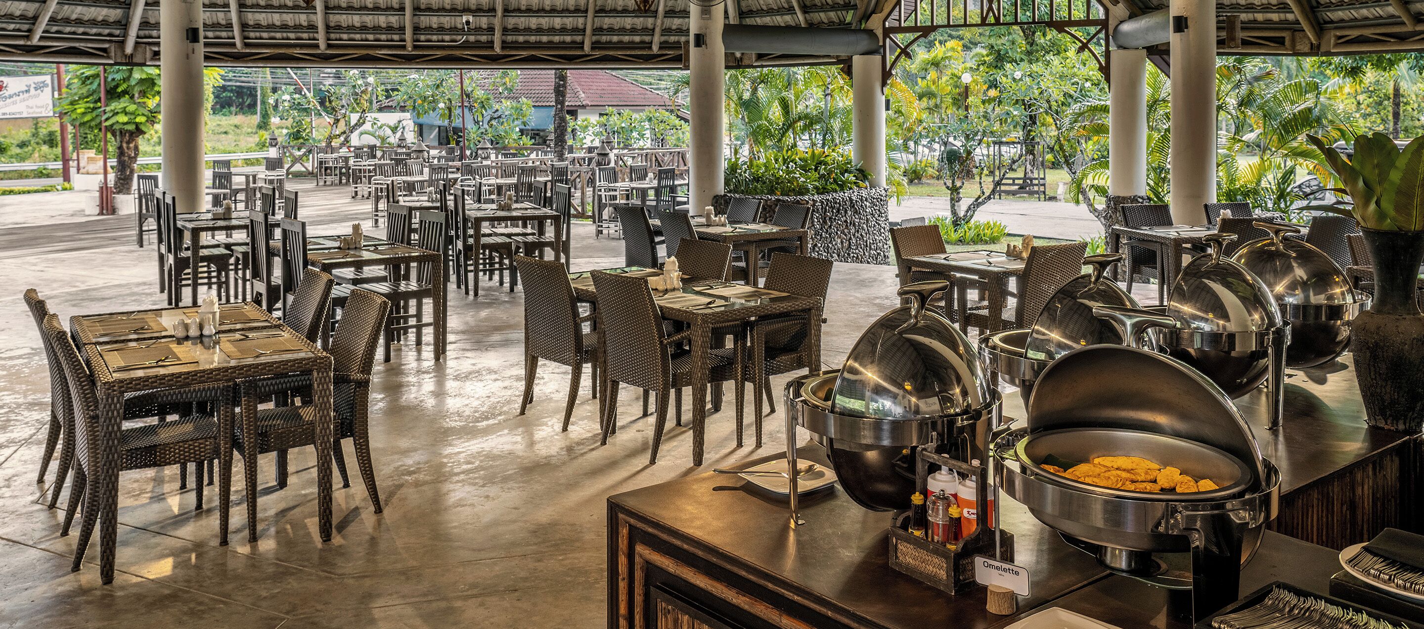 A covered outdoor dining area with multiple tables and chairs, a buffet station with food containers, and a view of the surrounding greenery.
