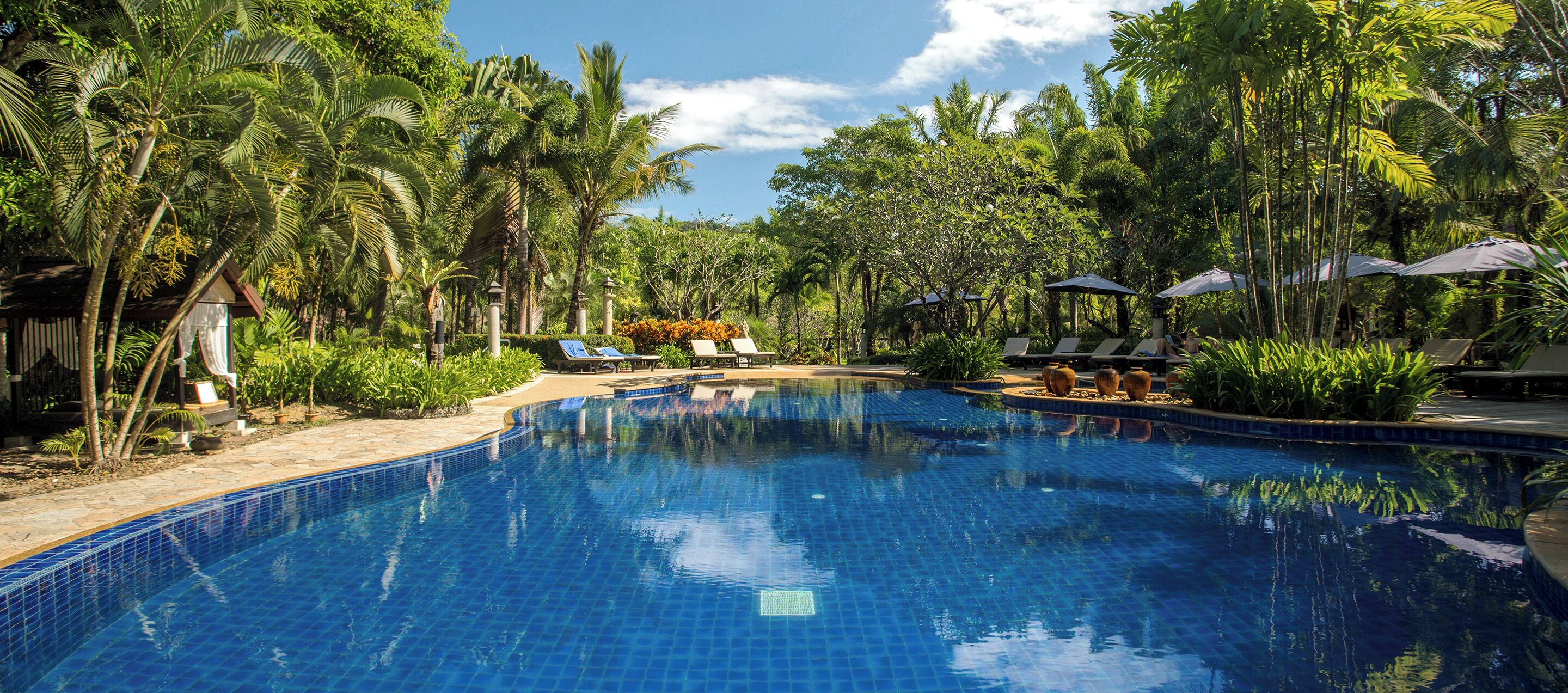 A clear blue swimming pool surrounded by lush greenery and palm trees, with lounge chairs and umbrellas for relaxation.