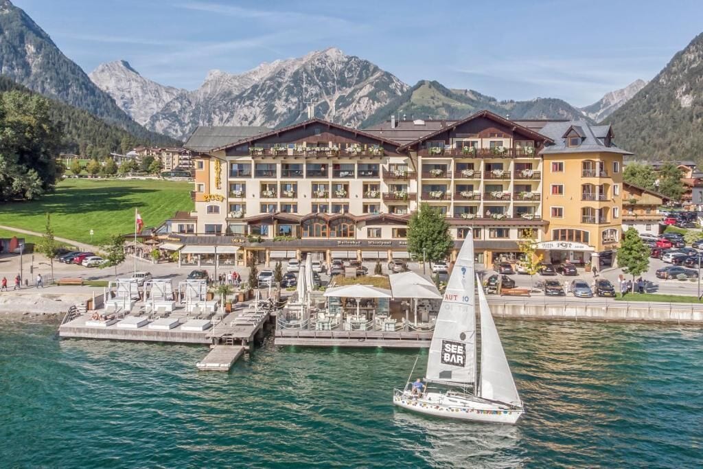 A hotel with multiple balconies overlooking a lake, a sailboat docked in front, and mountains in the background.