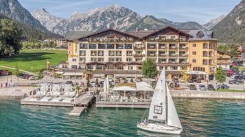 A hotel with multiple balconies overlooking a lake, a sailboat docked in front, and mountains in the background.