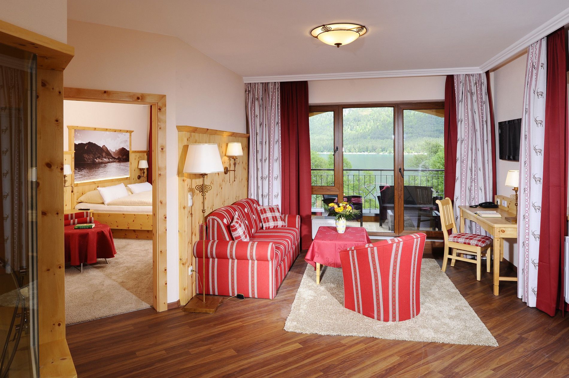 Habitación de hotel con una cama de madera, un sofá de rayas rojas, una mesa de comedor con sillas y un balcón con vistas a un lago.