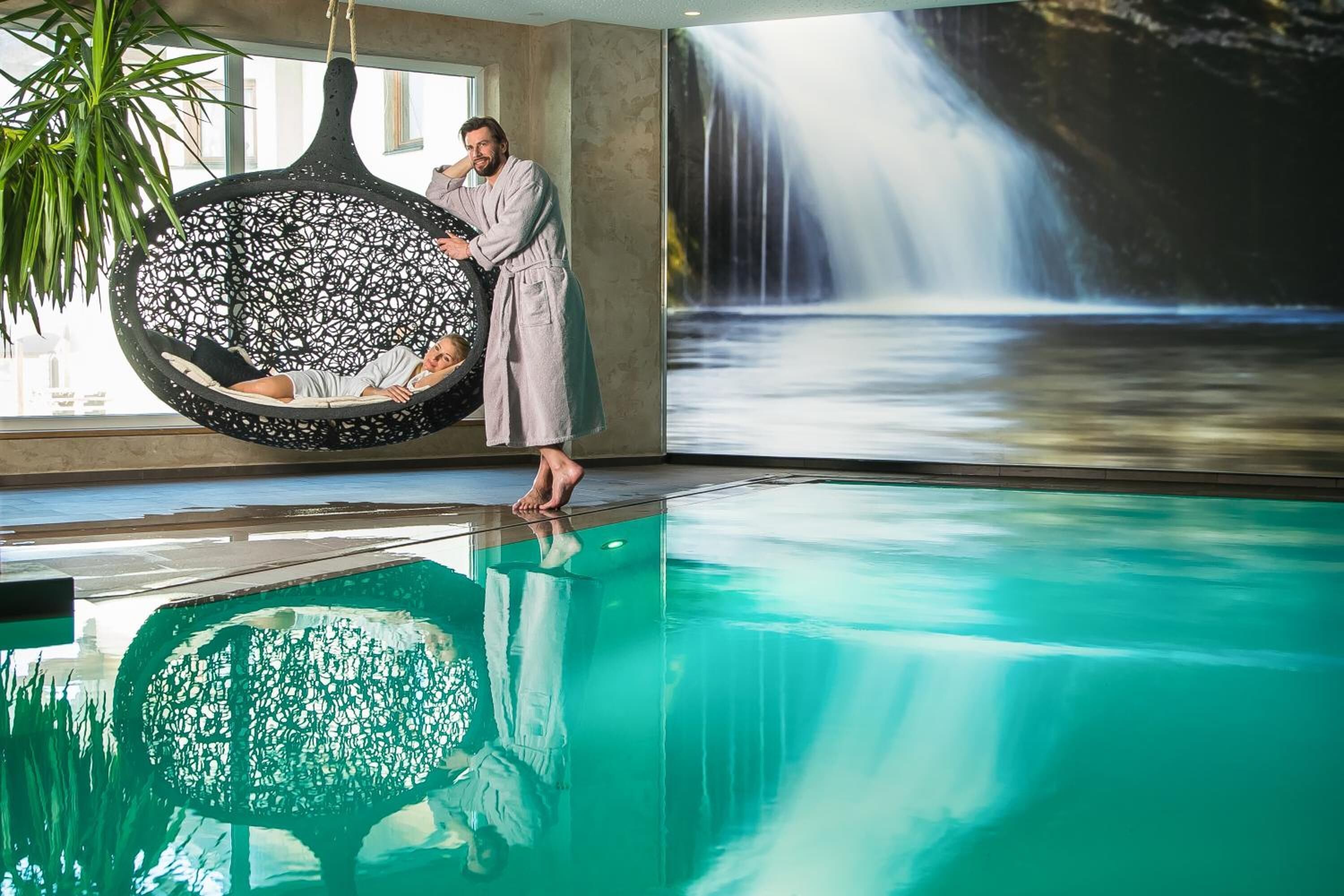 A man in a robe standing by a large indoor pool with a hanging chair and a waterfall mural.