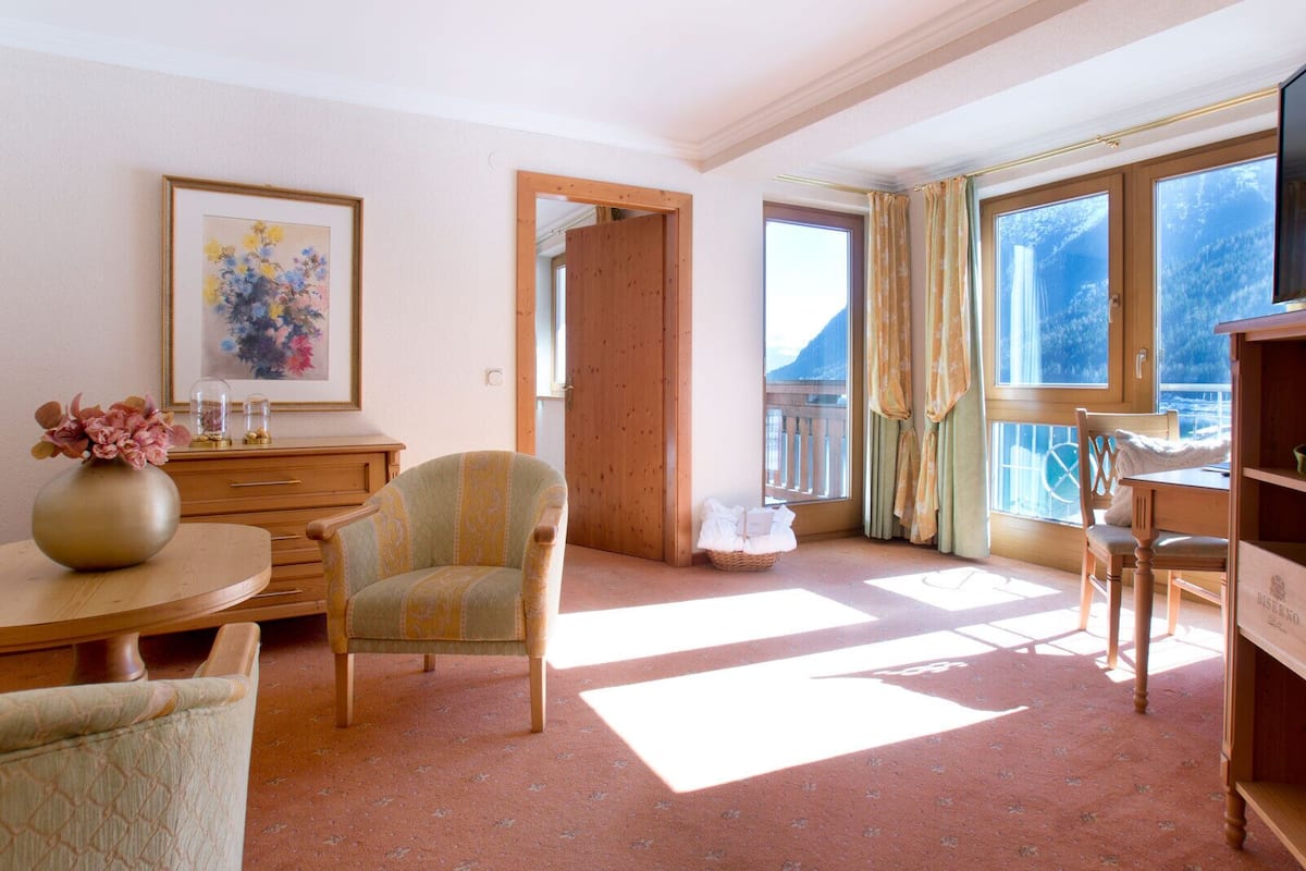Ein gemütliches Zimmer mit einem Holzstuhl, einem kleinen Tisch mit einer Vase, einem Schminktisch und einem Fenster mit Vorhängen.