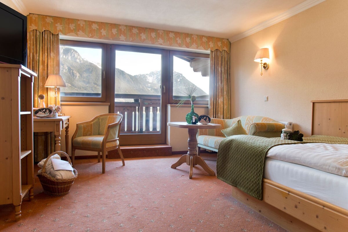 Ein Hotelzimmer mit Bett, Sessel, kleinem Tisch, Lampe, Fernseher und einem Fenster mit Bergblick.