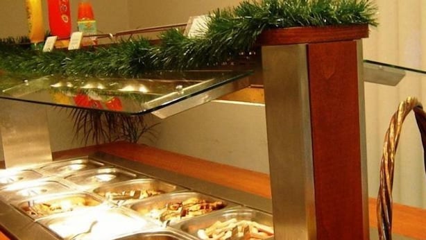 Un bufé con diversos alimentos en recipientes metálicos, una zona de servicio con cubierta de vidrio y una cesta de naranjas.