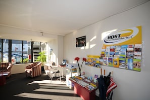 Ein Empfangsbereich mit Schreibtisch, Stühlen, einem Computer und einer Wand mit dem Schild 'HOST Accommodation' sowie verschiedenen Informationsbroschüren.