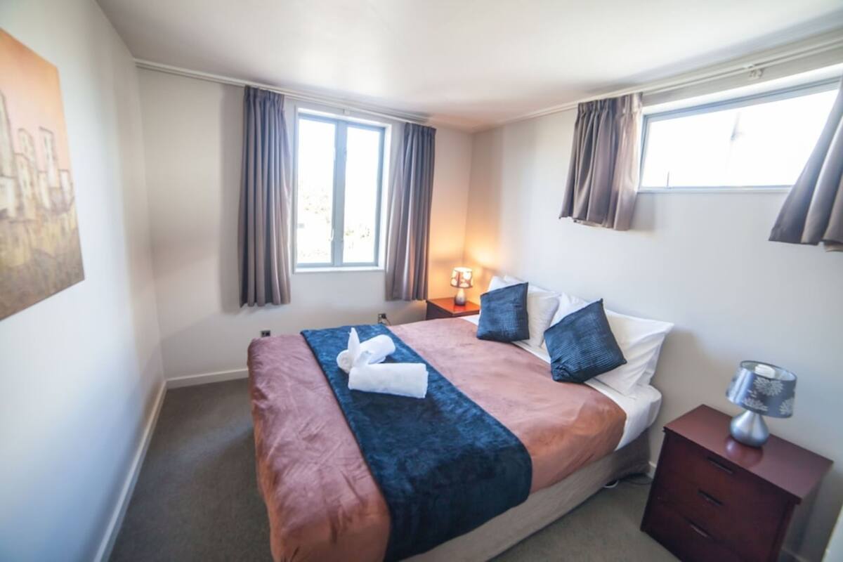 Habitación de hotel con cama, mesita de noche, lámpara y ventana con cortinas.