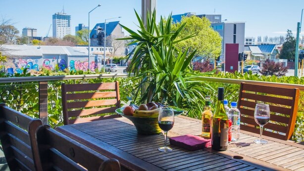 Een houten tafel met een fruitschaal, wijnglazen en flessen, op een balkon met uitzicht op gebouwen en groen.