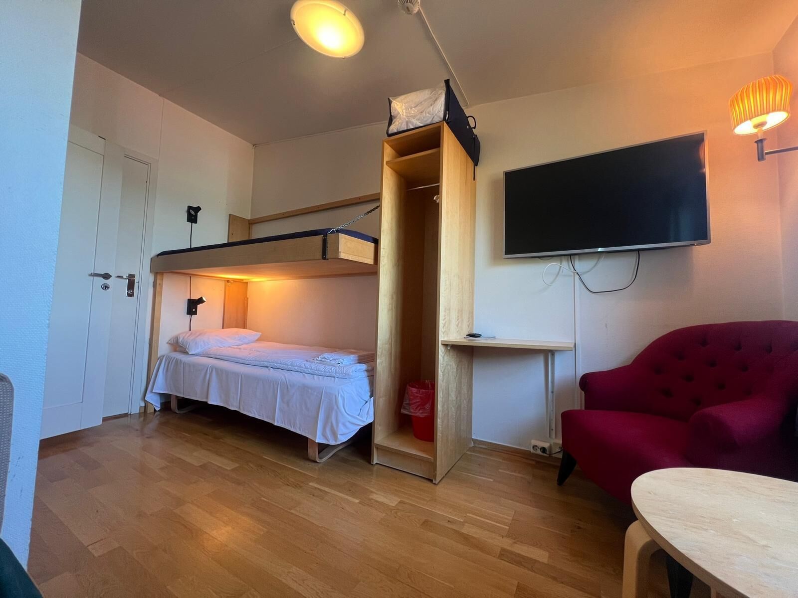 A compact room with a bunk bed, a red armchair, a small table, and a TV mounted on the wall.
