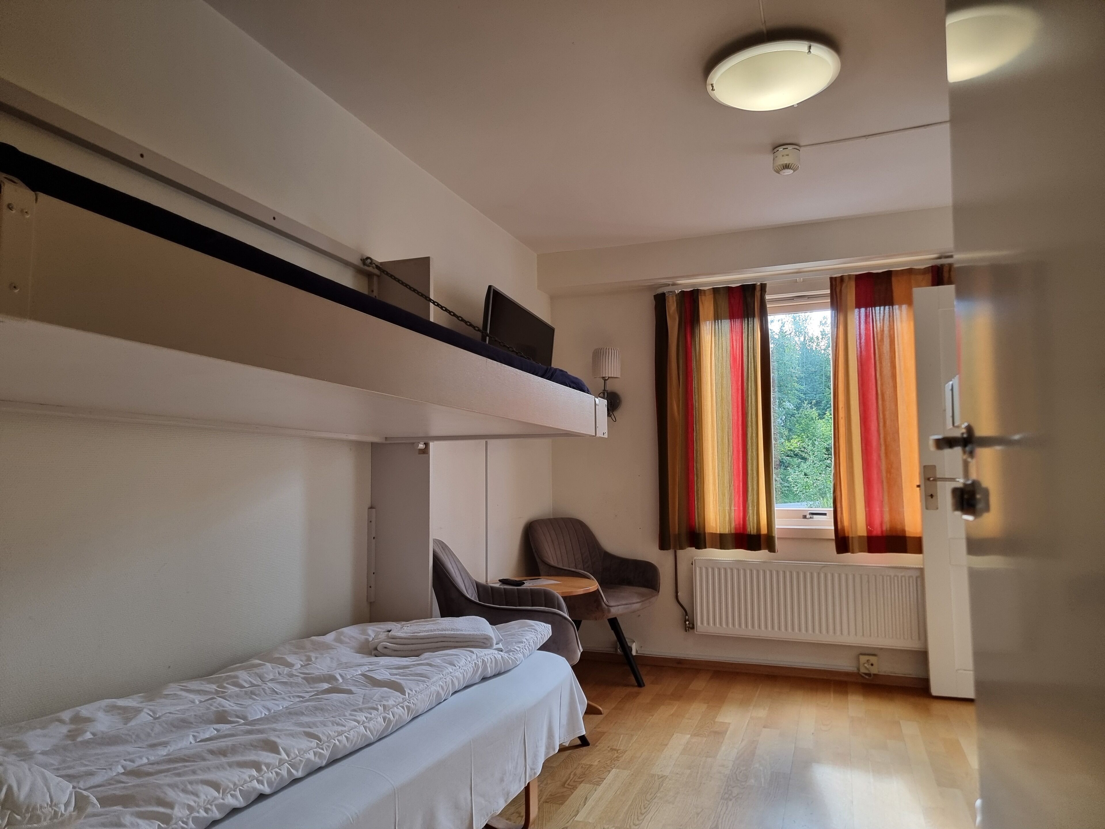 Una habitación con una cama alta, una cama individual, una silla, un radiador y una ventana con cortinas.